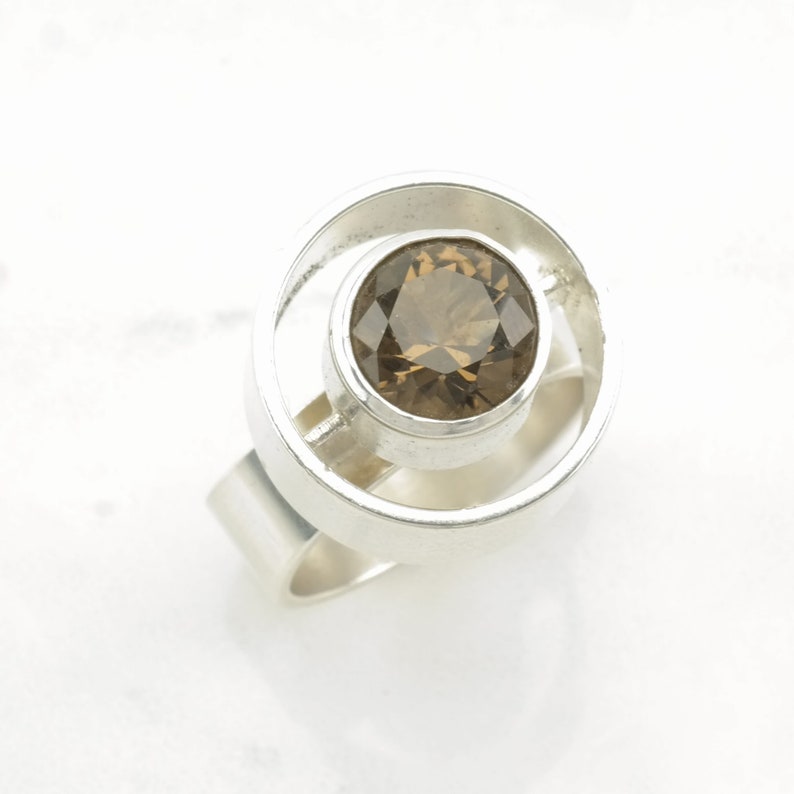 Peut inclure: Une bague en argent avec une pierre pr&eacute;cieuse ronde de couleur brune sertie dans un cadre circulaire. La bague a une bande simple.