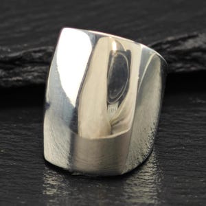 Puede incluir: Un anillo ancho de plata pulida con una superficie lisa y reflectante. La banda presenta un diseño curvo y cónico que se envuelve alrededor del dedo, mostrando un acabado metálico brillante que captura la luz en su exterior de metal sólido.