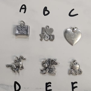 Op de afbeelding: Een verzameling zilverkleurige bedels, waaronder een koffer met de tekst "Paris" en "NYC", een "I Love You" bedel, een hartvormige bedel met de tekst "King of Hearts", een paard, een engel en een appel met "ABC".