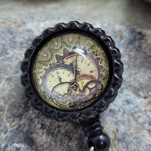 May include: Black retractable badge holder with a round, black and gold patterned button featuring a clock face design.