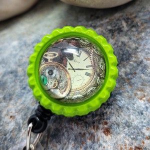 May include: A green retractable badge holder with a round, clear, plastic cover featuring a vintage clock face with Roman numerals. The clock face is surrounded by gears and other steampunk-inspired elements.