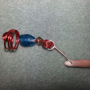 May include: A red wire wrapped around a blue glass bead with a brown bead and a silver wire attached to a brown shell.