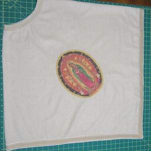 May include: A light beige t-shirt with a crew neck and a decorative image of Our Lady of Guadalupe. The image is oval-shaped with a gold border and features a depiction of the Virgin Mary in pink and green. The shirt has a beige trim.