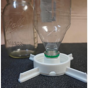 Pode incluir: Uma garrafa de plástico transparente é inserida em um dispositivo branco em forma de funil com quatro braços estabilizadores. Um frasco de vidro Mason está no fundo. A garrafa tem um anel verde ao redor do pescoço.