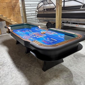 May include: A large, black craps table with a blue playing surface and a green padded rail. The table has a wooden trim and is set in a garage. The table has the words "PASS LINE" and other text.