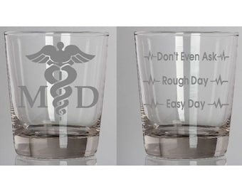 May include: Two clear glass tumblers with a medical theme. One tumbler has a caduceus symbol and the letters "MD". The other tumbler has the text "Don't Even Ask", "Rough Day", and "Easy Day" with a heartbeat symbol next to each phrase.