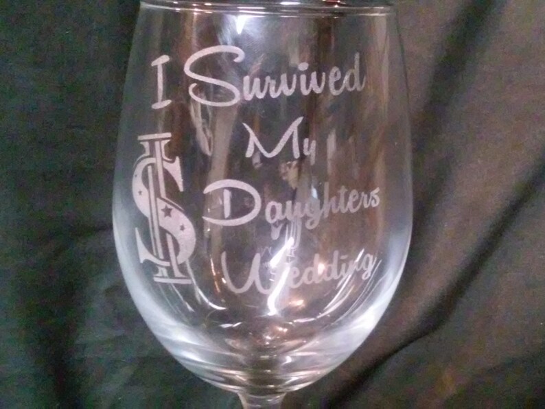 May include: A clear glass wine glass with the text "I Survived My Daughters Wedding" etched on the side. The text is in a cursive font and includes a stylized dollar sign.