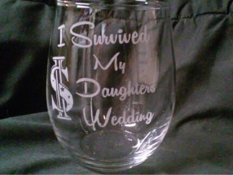 May include: A clear glass wine glass with the text "I Survived My Daughters Wedding" etched on the side. The text is in a white font and includes a dollar sign graphic.