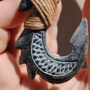 May include: A black carved fish hook pendant with a detailed design. The hook is tied with a brown cord.