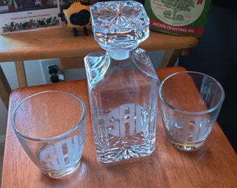 Pode incluir: Uma garrafa de vidro transparente com base quadrada e uma rolha de cristal. Dois copos transparentes com iniciais gravadas estão sobre uma superfície de madeira. A garrafa e os copos são monogramados com as iniciais "SAL".