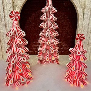 May include: Three decorative Christmas trees with red and white striped designs, resembling candy canes. Each tree has a peppermint candy on top and is illuminated with a warm glow. The trees are set against a brick background.