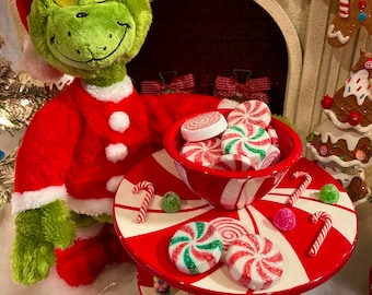 Könnte beinhalten: Ein grünes Grinch-Plüschtier, das einen roten Weihnachtsmann-Anzug trägt, steht neben einem roten und weißen, mit Zuckerstangen verzierten, mehrstöckigen Serviertablett mit Pfefferminzbonbons und Zuckerstangen. Das Tablett steht auf einer weißen Oberfläche mit grünen, orangefarbenen und violetten Wirbelornamenten.