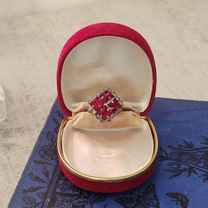 May include: A red velvet ring box with a gold trim sits on a blue book. Inside the box is a red gemstone ring with a square shape and smaller clear gemstones around the edges.