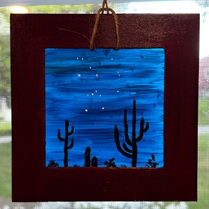 May include: A framed painting of a desert night scene with a blue sky, white stars, and three black silhouette cacti. The frame is dark red wood.