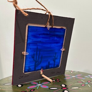 May include: A framed artwork depicting a blue desert night sky with silhouetted cacti. The frame is made of wood and has a dark gray finish. The artwork is titled "Orion" and is signed "Jen Lowe".
