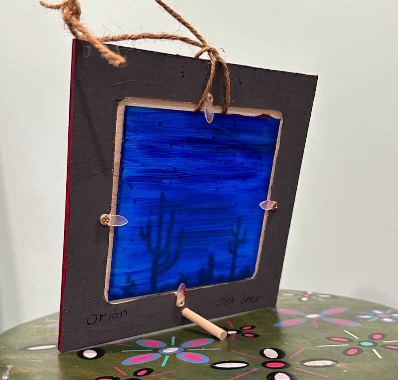 May include: A framed artwork depicting a blue desert night sky with silhouetted cacti. The frame is made of wood and has a dark gray finish. The artwork is titled "Orion" and is signed "Jen Lowe".