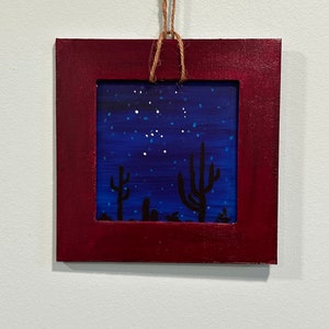 May include: A framed painting of a desert night scene with a dark blue sky filled with white stars and two black silhouettes of cacti in the foreground.