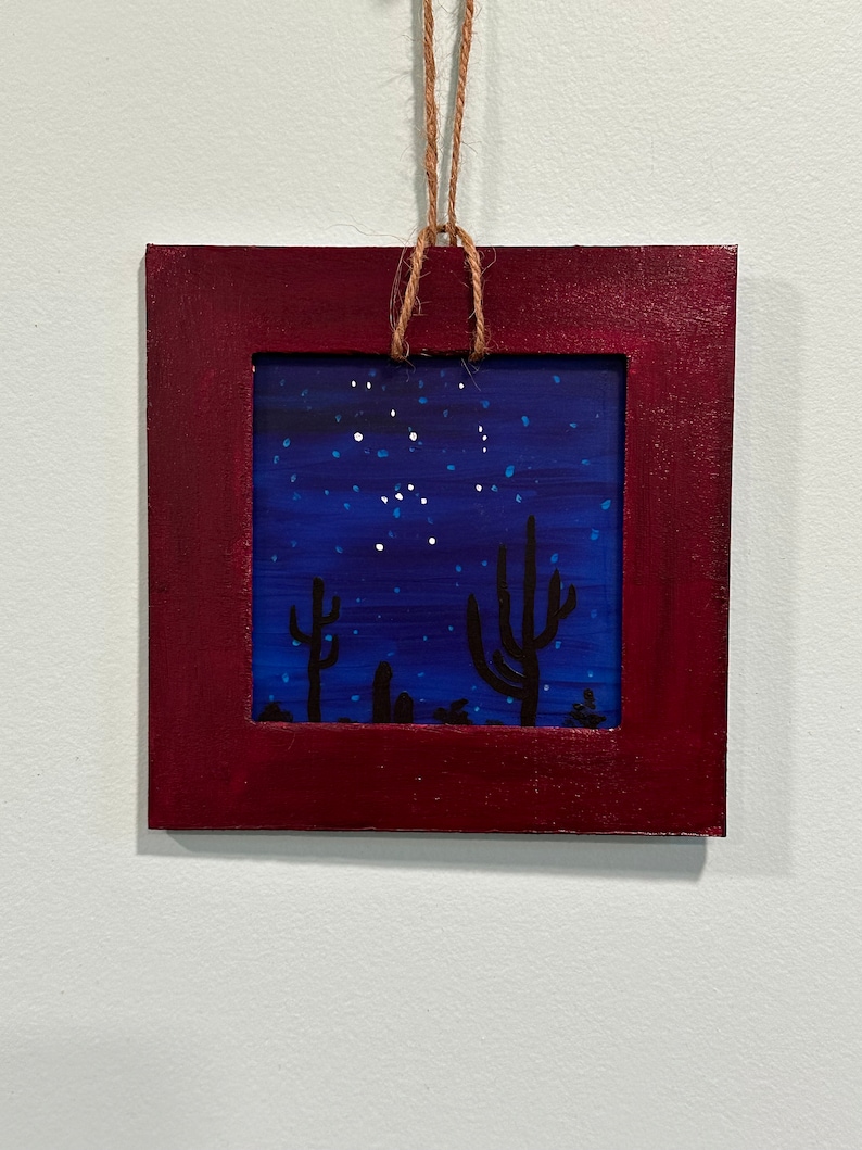 May include: A framed painting of a desert night scene with a dark blue sky filled with white stars and two black silhouettes of cacti in the foreground.