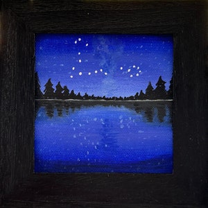 May include: A framed painting of a dark blue night sky with white stars forming a constellation above a silhouette of a forest reflected in a still lake.