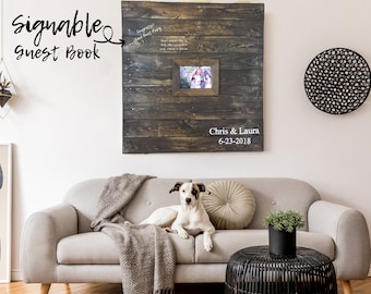May include: A rustic wooden sign with a frame for a photo, with the text "Chris & Laura 6-23-2018" written on it. The sign is hanging on a white wall above a gray couch with a white dog lying on it. There is a black wicker side table next to the couch.