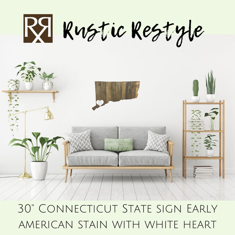 May include: 30" Connecticut state sign with a white heart, made of reclaimed wood with an early American stain. The sign is displayed on a white wall in a living room setting with a grey sofa, a wooden shelf, and plants.