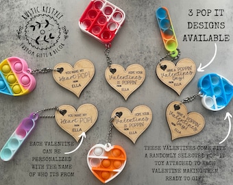 Puede incluir: Regalos de San Valentín con etiquetas de madera en forma de corazón y juguetes pop-it. Las etiquetas tienen frases como "You Make My Heart POP!". El texto de la imagen dice "3 POP IT DESIGNS AVAILABLE". Cada regalo puede ser personalizado.