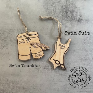 May include: Two wooden ornaments shaped like swim trunks and a swimsuit. The swim trunks ornament has the name "Cody" and the year "2023" engraved on it. The swimsuit ornament has the name "Michelle" and the year "2023" engraved on it. Both ornaments have a pair of goggles engraved on them. The ornaments are hanging from twine. The text "Swim Trunks" is below the swim trunks ornament and the text "Swim Suit" is above the swimsuit ornament. The text "Rustic Restyle Custom Gifts & Decor" is below the swimsuit ornament.