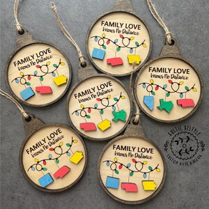 May include: Five circular wooden ornaments with twine hangers. Each features the text "FAMILY LOVE Knows No Distance" above a string of colourful Christmas lights and various U.S. state shapes in red, blue, yellow, and green.