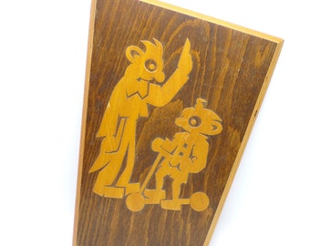 May include: A wooden wall hanging with a carved design of two figures. The figures are stylized and appear to be wearing clothes. The larger figure is holding up a hand, while the smaller figure is riding a scooter. The background is a dark brown wood.
