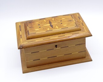 May include: A wooden box with a decorative inlay design on the lid. The box is made of light wood with dark wood accents. The box has a keyhole on the front.