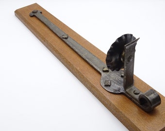 May include: A long, thin, metal hinge with a decorative wheel at one end. The hinge is mounted on a wooden board.