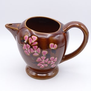 Op de afbeelding: Een bruine keramische kan met een handvat en een tuit. De kan is versierd met roze bloemen.