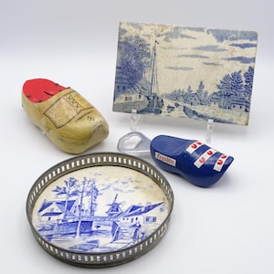 Könnte beinhalten: Eine Sammlung niederländischer Souvenirs, darunter ein Holzschuh-Sparschwein, eine blau-weiße Fliese mit einer Kanalszene, ein Flaschenöffner in Holzschuhform und ein dekoratives Tablett mit blau-weißem Windmühlen-Design.