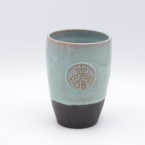 Puede incluir: Una taza de cerámica azul claro con un borde negro y un logotipo circular marrón que dice "1937-1962 JB" con una corona sobre las letras.