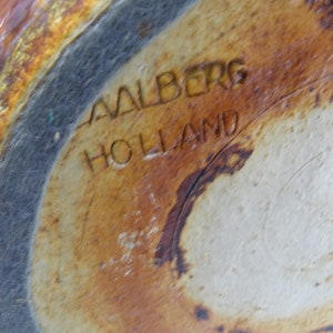 May include: Close-up of the base of a ceramic piece with the text "AALBERG HOLLAND" etched into the surface. The ceramic has a mottled, earthy color palette of brown, tan, and gray.