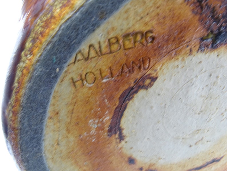 May include: Close-up of the base of a ceramic piece with the text "AALBERG HOLLAND" etched into the surface. The ceramic has a mottled, earthy color palette of brown, tan, and gray.