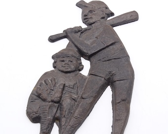 May include: A bronze-colored metal sculpture depicting two baseball players. One player is holding a bat over their shoulder, while the other is in a catcher's stance. The sculpture has a textured surface and is set against a white background.