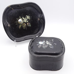 May include: A black, vintage jewelry box and matching tray. Both items feature a floral design with iridescent accents and a central pearl-like embellishment. The tray has a unique, curved shape.