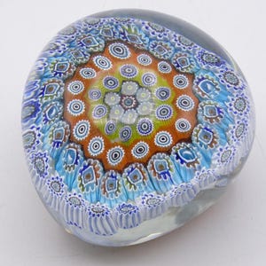 May include: A clear glass paperweight with a colorful millefiori design. The center features a circular pattern of orange, yellow, and white, surrounded by blue and white floral motifs. The paperweight is approximately 3 inches in diameter.