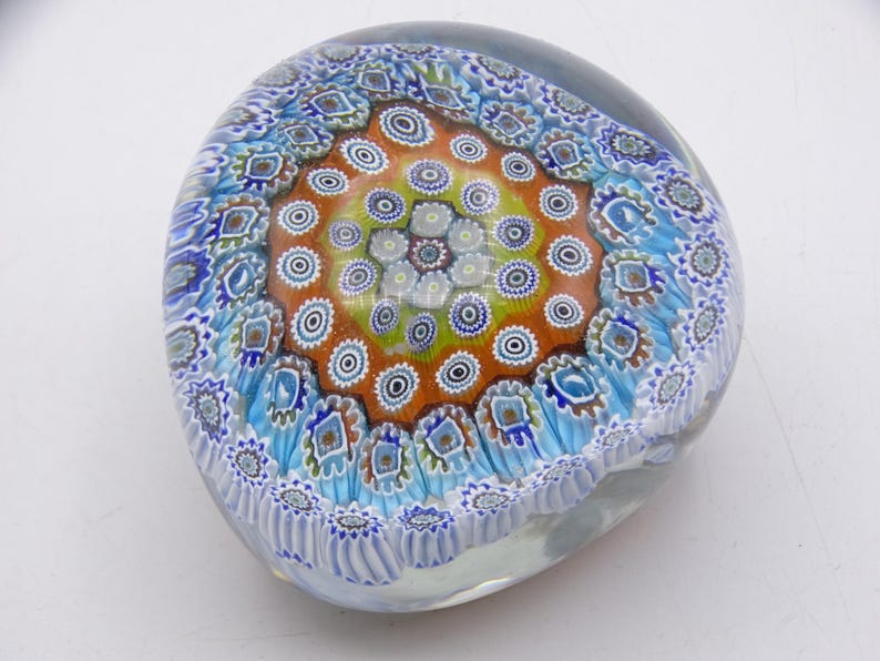 May include: A clear glass paperweight with a colorful millefiori design. The center features a circular pattern of orange, yellow, and white, surrounded by blue and white floral motifs. The paperweight is approximately 3 inches in diameter.