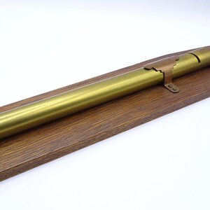 K&ouml;nnte beinhalten: Ein goldfarbenes Metallrohr mit einer roten und kupferfarbenen Endkappe ist auf einem Holzsockel montiert. Das Rohr ist etwa 30 cm lang.