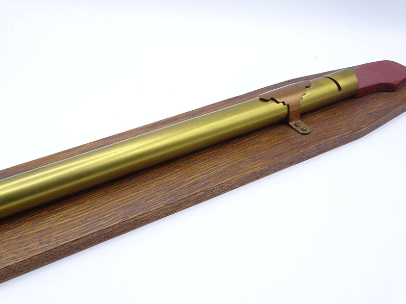 K&ouml;nnte beinhalten: Ein goldfarbenes Metallrohr mit einer roten und kupferfarbenen Endkappe ist auf einem Holzsockel montiert. Das Rohr ist etwa 30 cm lang.