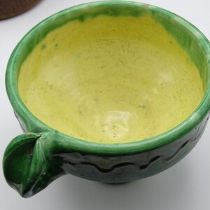 May include: A handmade ceramic bowl with a bright yellow interior and a green rim and handle. The bowl has a wavy design around the exterior and is likely used for serving food. The bowl is a unique piece of kitchenware.