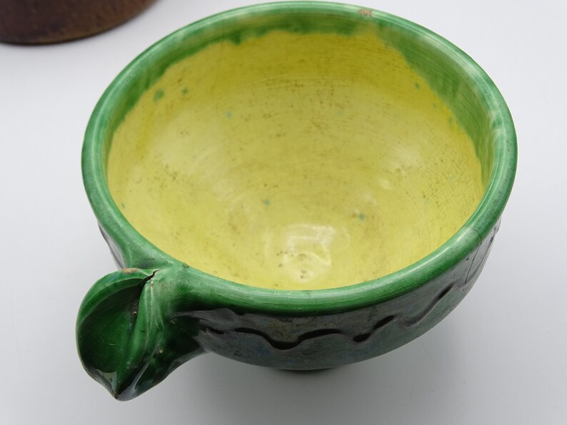 May include: A handmade ceramic bowl with a bright yellow interior and a green rim and handle. The bowl has a wavy design around the exterior and is likely used for serving food. The bowl is a unique piece of kitchenware.
