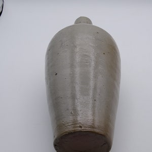 May include: A tall, antique stoneware jug with a rounded body and a narrow neck. The jug is a muted gray-brown color with a darker brown base. The surface has a slightly glossy finish and visible imperfections.