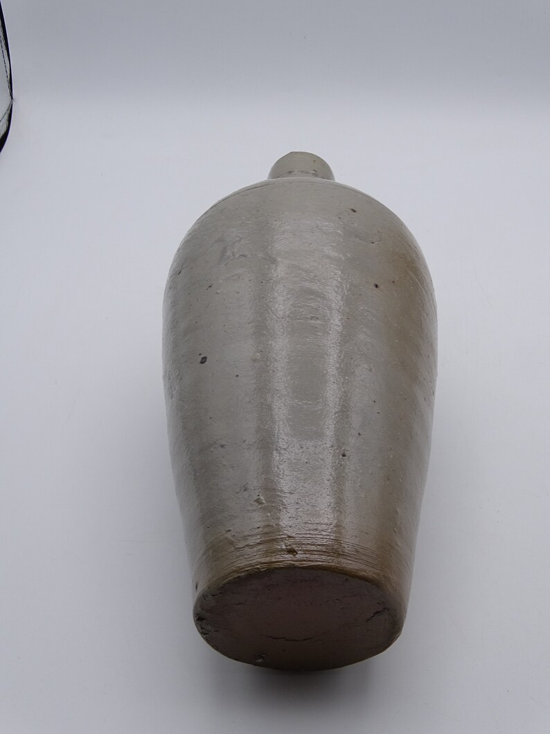 May include: A tall, antique stoneware jug with a rounded body and a narrow neck. The jug is a muted gray-brown color with a darker brown base. The surface has a slightly glossy finish and visible imperfections.