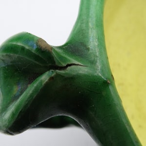 May include: Close-up of a ceramic handle, part of a larger object. The handle is a vibrant green with a glossy finish, showing signs of wear. A crack is visible. The background is a soft yellow.