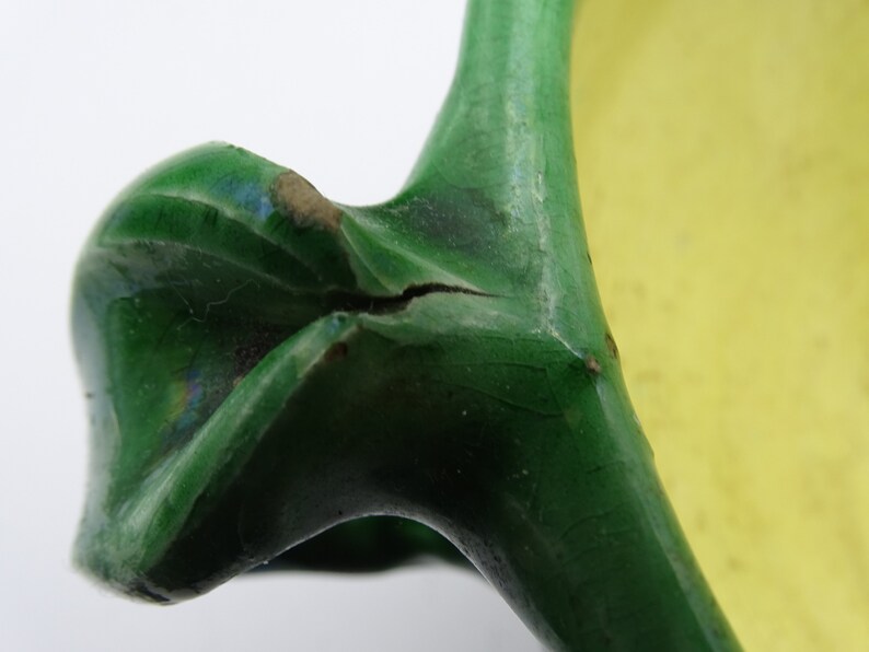 May include: Close-up of a ceramic handle, part of a larger object. The handle is a vibrant green with a glossy finish, showing signs of wear. A crack is visible. The background is a soft yellow.