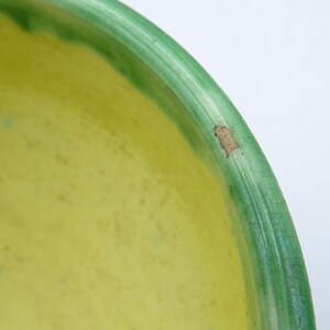 May include: Close-up of a ceramic bowl with a bright yellow interior and a green rim. The green rim has a darker green border. There is a small area of damage on the rim. The bowl is on a white background.