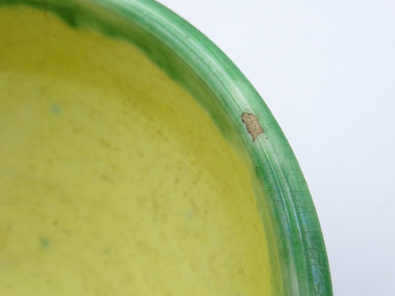 May include: Close-up of a ceramic bowl with a bright yellow interior and a green rim. The green rim has a darker green border. There is a small area of damage on the rim. The bowl is on a white background.